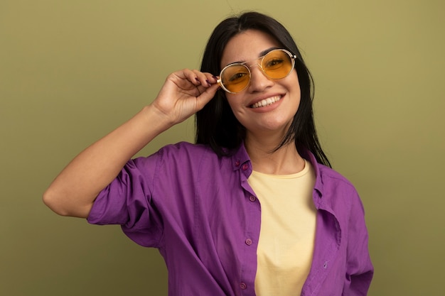 Sonriente niña caucásica morena bonita mira a la cámara a través de gafas de sol en verde oliva