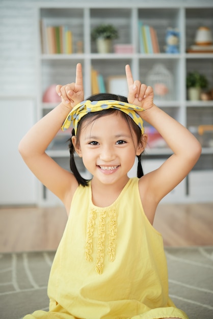 Sonriente niña china sentada en casa y haciendo gesto de cuernos de vaca