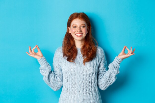 Sonriente niña confiada con cabello rojo siendo paciente, tomados de la mano en zen, pose de meditación y mirando a la cámara, practica yoga, de pie tranquilo contra el fondo azul.