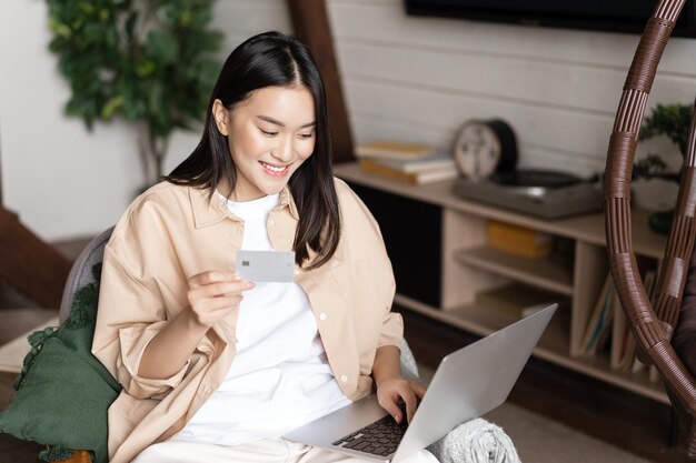 Sonriente niña coreana comprando en línea desde casa, compras en una computadora portátil y sosteniendo una tarjeta de crédito