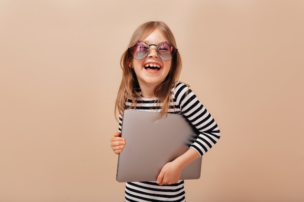 Sonriente niña emocionada con gafas y camisa despojada riendo y sosteniendo la computadora portátil