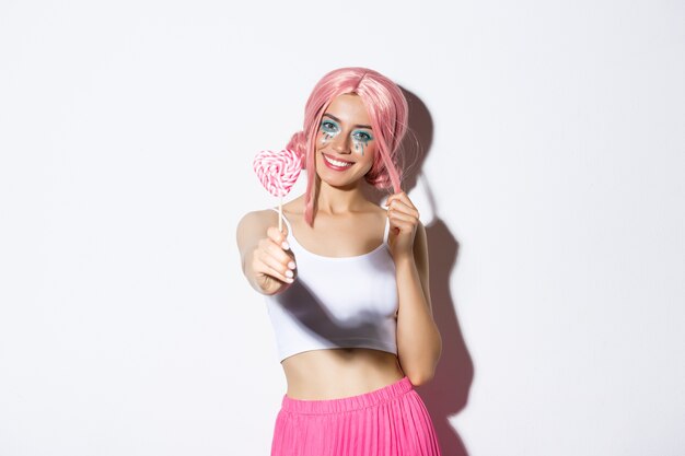 Sonriente niña encantadora con peluca rosa que le da dulces, celebrando halloween, truco o trato.