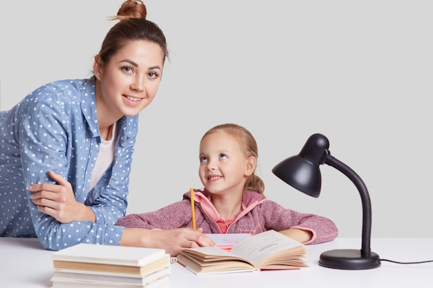 La sonriente niña encantadora se sienta a la mesa, hace la tarea junto con su madre, trata de escribir la composición, mira alegremente, usa una lámpara de lectura para una buena visión, aislada en la pared blanca