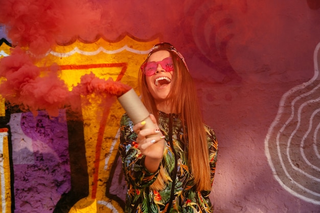 Sonriente niña feliz en gafas de sol con bomba de humo rojo, de pie cerca de la pared con graffiti.