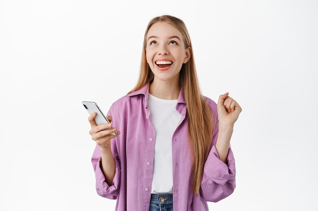 Sonriente niña feliz mirando hacia arriba aliviada después de ganar en el teléfono móvil, sosteniendo el teléfono inteligente y regocijándose, celebrando el logro en la aplicación, de pie sobre una pared blanca.