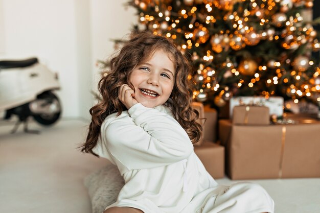 Sonriente niña feliz con pelo rizado vistiendo suéter de punto blanco posando con una sonrisa feliz mientras está sentado del árbol de Navidad