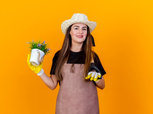 Sonriente niña hermosa jardinera vestida con uniforme y sombrero de jardinería con guantes poniendo la pala en el hombro y sosteniendo la flor en maceta aislado sobre fondo naranja