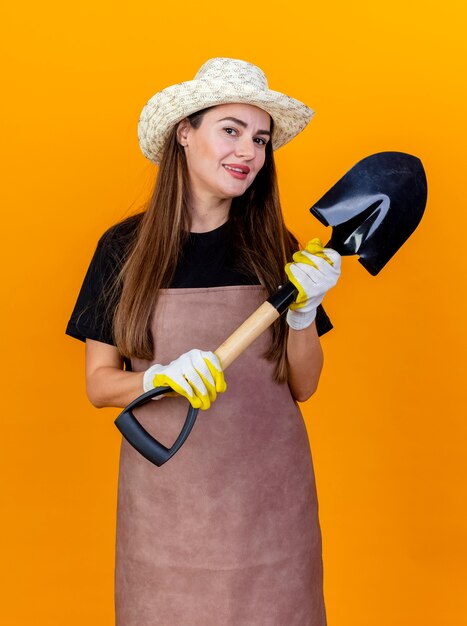 Sonriente niña hermosa jardinera vestida con uniforme y sombrero de jardinería con guantes sosteniendo pala aislado en naranja