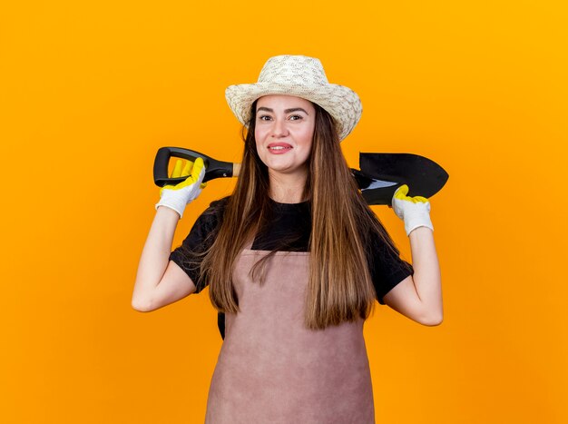 Sonriente niña hermosa jardinera vestida con uniforme y sombrero de jardinería con guantes sosteniendo la pala detrás del cuello aislado sobre fondo naranja