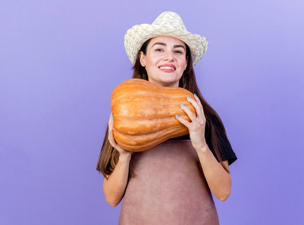 Sonriente niña hermosa jardinero en uniforme con sombrero de jardinería con calabaza aislado en azul