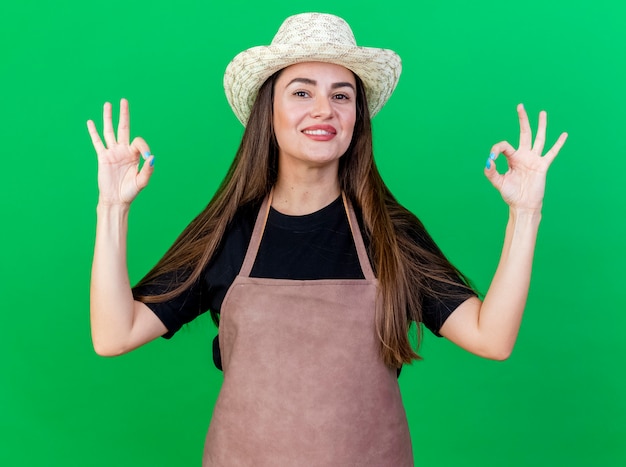 Sonriente niña hermosa jardinero en uniforme con sombrero de jardinería mostrando gestos okey aislados sobre fondo verde