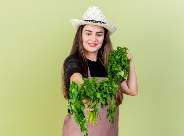 Sonriente niña hermosa jardinero en uniforme con sombrero de jardinería sosteniendo cilantro en cámara aislada sobre fondo verde oliva