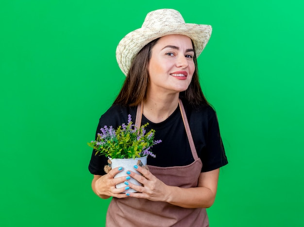 Foto gratuita sonriente niña hermosa jardinero en uniforme con sombrero de jardinería sosteniendo flor en maceta aislado en verde