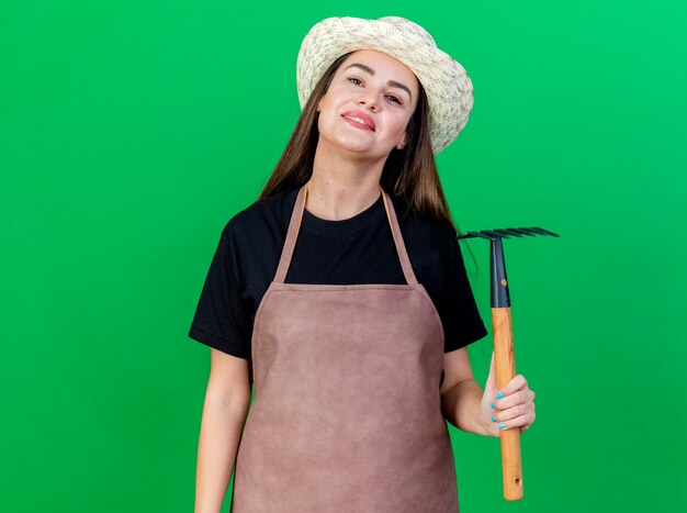 Sonriente niña hermosa jardinero en uniforme con sombrero de jardinería sosteniendo rastrillo aislado sobre fondo verde