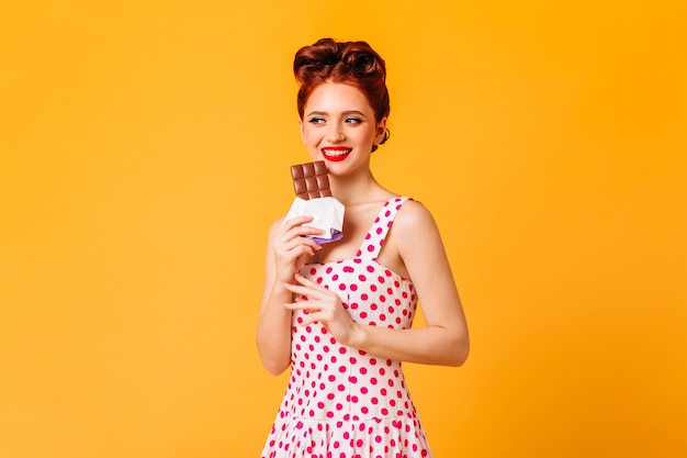 Sonriente niña de jengibre comiendo chocolate. Disparo de estudio de mujer pinup en vestido de lunares aislado en espacio amarillo.