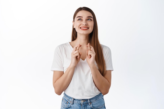 Sonriente niña linda pidiendo deseos, cruzando los dedos para tener suerte y mirando hacia arriba, rogando a Dios por buenos resultados positivos, anticipando noticias, de pie sobre fondo blanco.