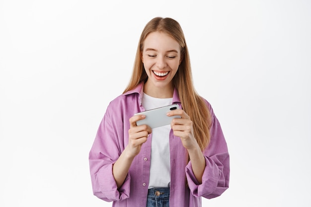 Sonriente niña moderna sosteniendo el teléfono inteligente en ambas manos, mirando la pantalla móvil horizontal, viendo videos en el teléfono o jugando videojuegos, de pie sobre una pared blanca