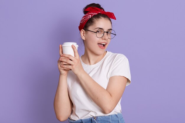 Sonriente niña morena en camiseta blanca casual sosteniendo una taza de café aislada sobre el espacio lila, mirando a la cámara, mantiene la boca abierta