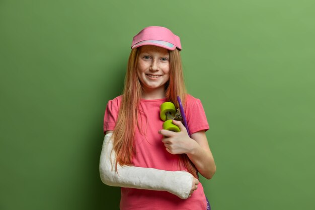 Sonriente niña pelirroja tuvo un accidente después de andar en patineta, usa yeso o yeso en el brazo roto, se mantiene feliz, se lesionó durante su deporte favorito, se para contra la pared verde. Niños, cuidado de la salud