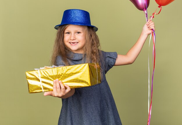 Sonriente niña rubia con gorro de fiesta azul sosteniendo globos de helio y caja de regalo aislado en la pared verde oliva con espacio de copia