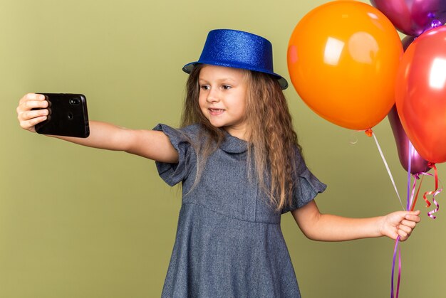 Sonriente niña rubia con gorro de fiesta azul sosteniendo globos de helio y teléfono tomando selfie aislado en la pared verde oliva con espacio de copia