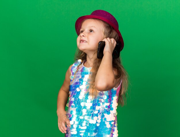 Sonriente niña rubia con gorro de fiesta púrpura hablando por teléfono mirando al lado aislado en la pared verde con espacio de copia