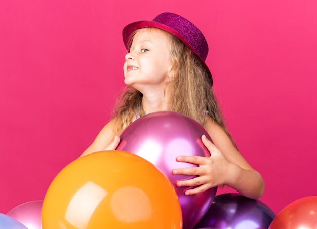 Sonriente niña rubia con gorro de fiesta púrpura de pie con globos de helio mirando al lado aislado en la pared rosa con espacio de copia