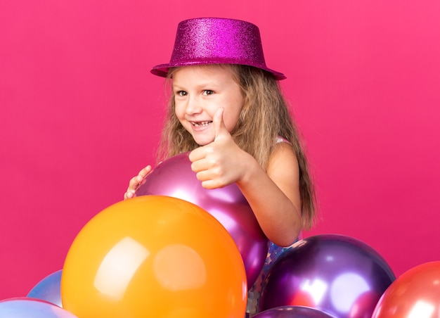 Sonriente niña rubia con gorro de fiesta púrpura de pie con globos de helio pulgar hacia arriba aislado en la pared rosa con espacio de copia