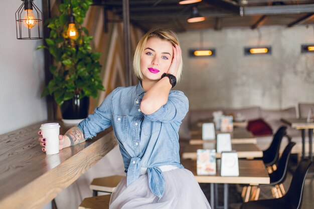 Sonriente niña rubia sentada en una cafetería tomando café y tocando su cabello. Hay sillas y mesas de madera en la cafetería.