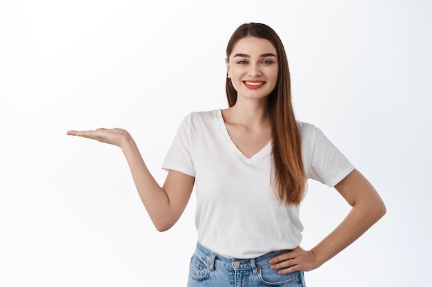 Sonriente niña sana estira la mano abierta y sostiene su logotipo, producto promocional, muestra un artículo en el espacio de la copia, recomienda comprar artículo, de pie sobre fondo blanco