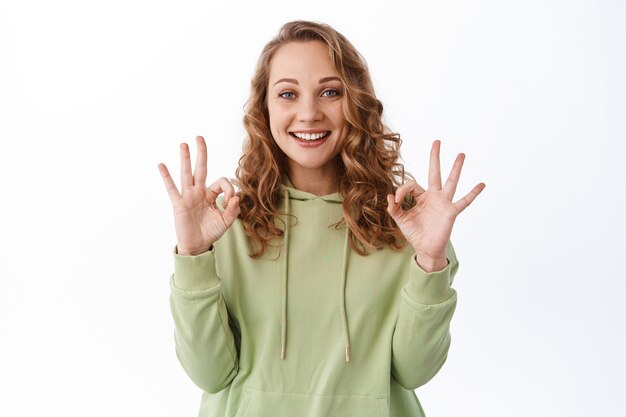 Sonriente niña satisfecha muestra signos de aprobación y asiente con la cabeza, elogia la buena elección, el producto perfecto, recomienda algo, pared blanca