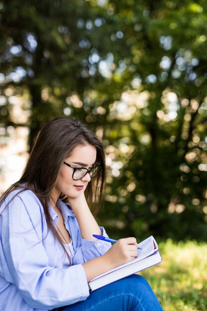 Sonriente niña seria de pelo oscuro en chaqueta de jeans y gafas escribir en un cuaderno en el parque