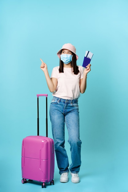 Sonriente niña turista viajero asiático en mascarilla de pie cerca de la maleta con dos boletos y pasaporte ...