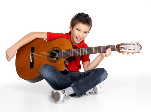 Sonriente niño caucásico está tocando la guitarra acústica aislado en blanco