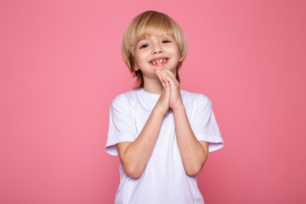 Sonriente niño rubio lindo dulce adorable en camiseta blanca en escritorio rosa