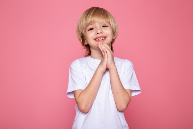 Sonriente niño rubio lindo dulce adorable en camiseta blanca en escritorio rosa