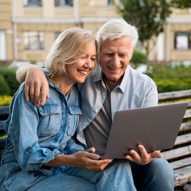 Sonriente pareja de ancianos sentados en un banco al aire libre con un portátil
