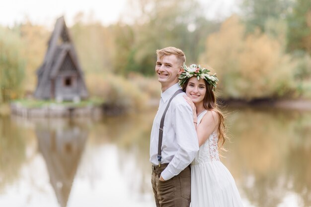 Sonriente pareja de enamorados se abraza cerca del pequeño lago, vestida con un acogedor atuendo de boda en el parque en otoño