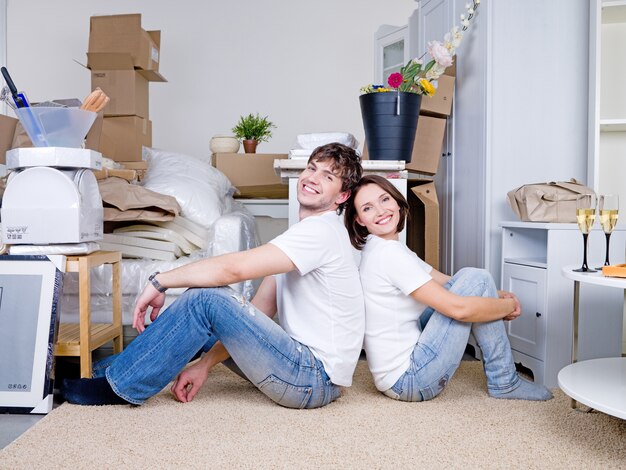 Sonriente pareja feliz sentados espalda con espalda en el suelo de la nueva casa