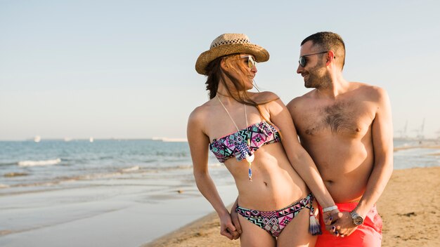 Sonriente pareja joven en traje de baño mirando mutuamente de pie en la playa