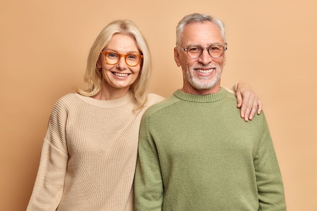 Sonriente pareja madura abrazar mira con alegría la pose de la cámara para el retrato de familia.Los niños felices vinieron a visitarlos. Usan lentes transparentes, puentes casuales aislados sobre una pared marrón.