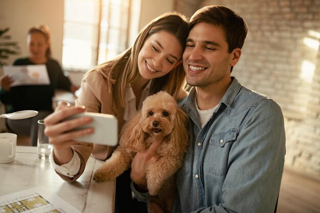 Sonriente pareja de negocios usando un teléfono inteligente mientras toma selfie con un caniche en la oficina