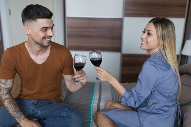 Sonriente pareja sentada en la cama brindando copas de vino