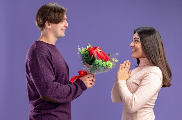 Sonriente de pie en la vista de perfil joven pareja en el día de San Valentín chico dando ramo a chica aislada sobre fondo azul.