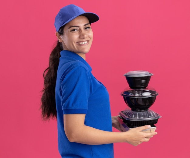 Sonriente de pie en la vista de perfil joven repartidora vistiendo uniforme con gorra sosteniendo contenedores de alimentos aislados en la pared rosa