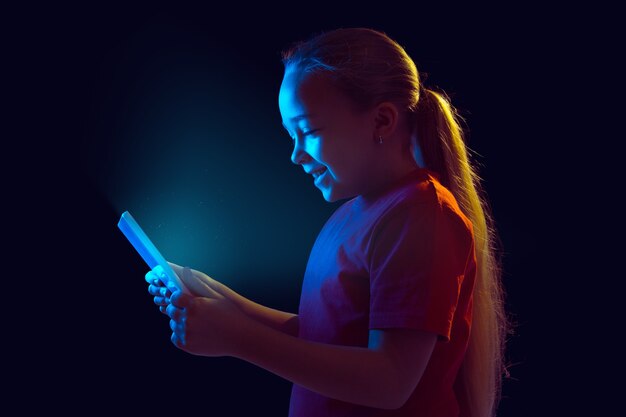 Sonriente. Retrato de niña caucásica aislado en la pared oscura en luz de neón. Modelo femenino hermoso que usa la tableta. Concepto de emociones humanas, expresión facial, ventas, publicidad, tecnología moderna, gadgets.