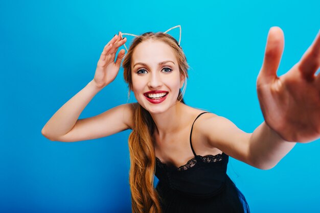 Sonriente rubia bonita con ojos azules posando, tomando selfie, disfrutando de la fiesta. Con vestido negro y diadema con orejas de gato.