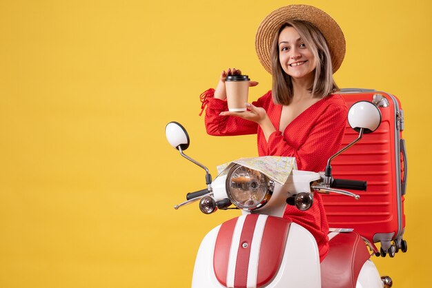 Sonriente señorita en vestido rojo sosteniendo una taza de café cerca del ciclomotor