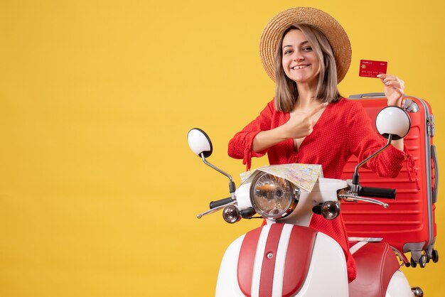 Sonriente señorita en vestido rojo con tarjeta de crédito cerca de ciclomotor