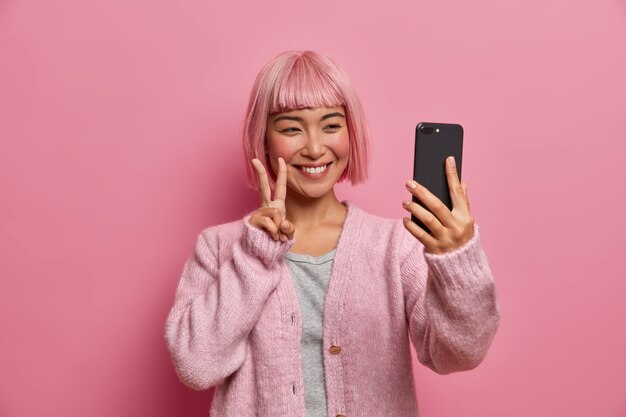 Sonriente y sincera mujer asiática hace un gesto de paz, da la bienvenida a un amigo durante la videoconferencia, se toma un selfie a través de un teléfono inteligente, está vestida con un jersey informal, tiene el cabello teñido a la moda,